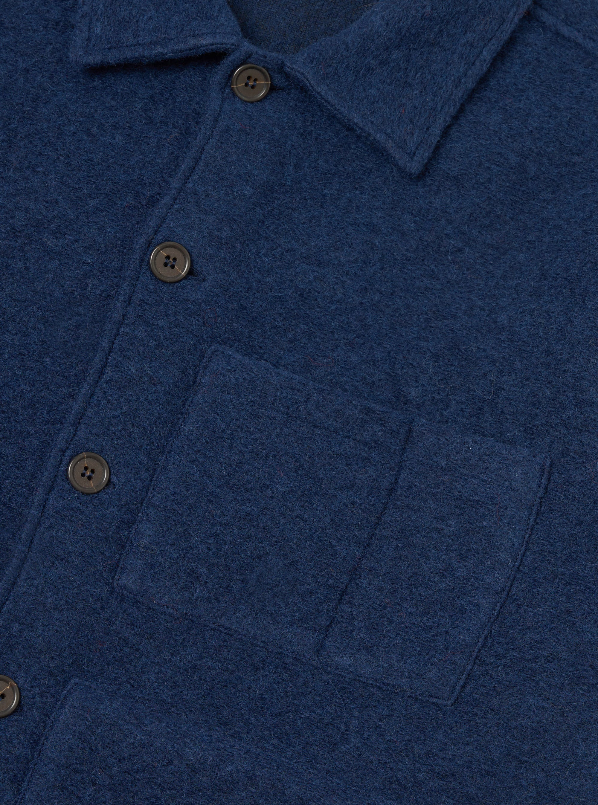 Close-up of an indigo wool fleece uniform jacket on a white studio backdrop. The brushed, textured fleece fabric is shown in detail, with a spread collar, four-hole dark horn-effect buttons on a front placket, and a large patch pocket on the chest.