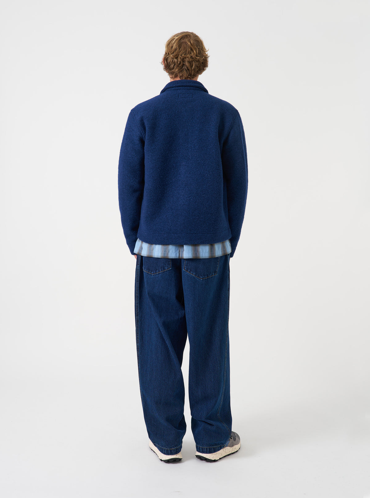Rear view of a model wearing the Indigo Wool Fleece Uniform Jacket against a light plain studio background. The model turns away to showcase the overall fit, straight hem, and deep indigo blue wool fleece material details.