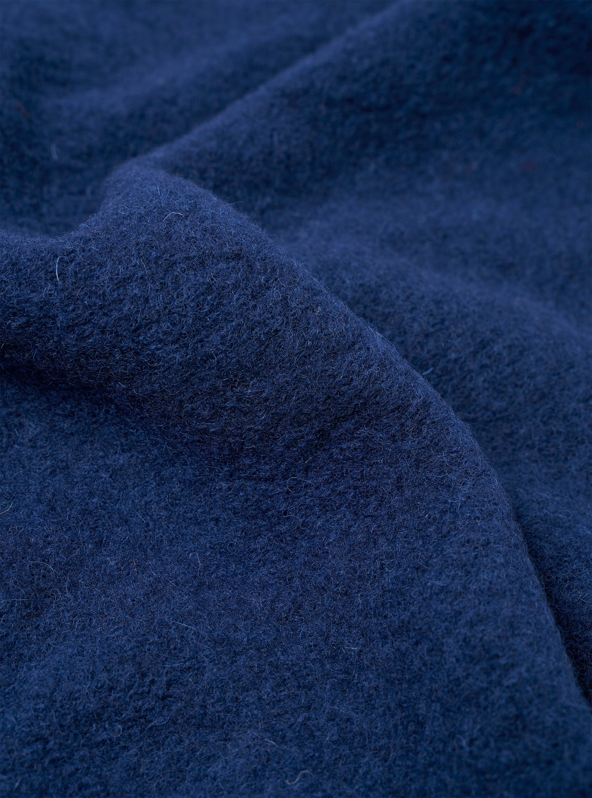 Close-up detail shot of the indigo wool fleece fabric used in the Uniform Jacket. The boiled or brushed wool surface shows a dense, lightly napped texture with fine curling fibres throughout. The deep navy-indigo colour is consistent across the draped folds visible in the frame.