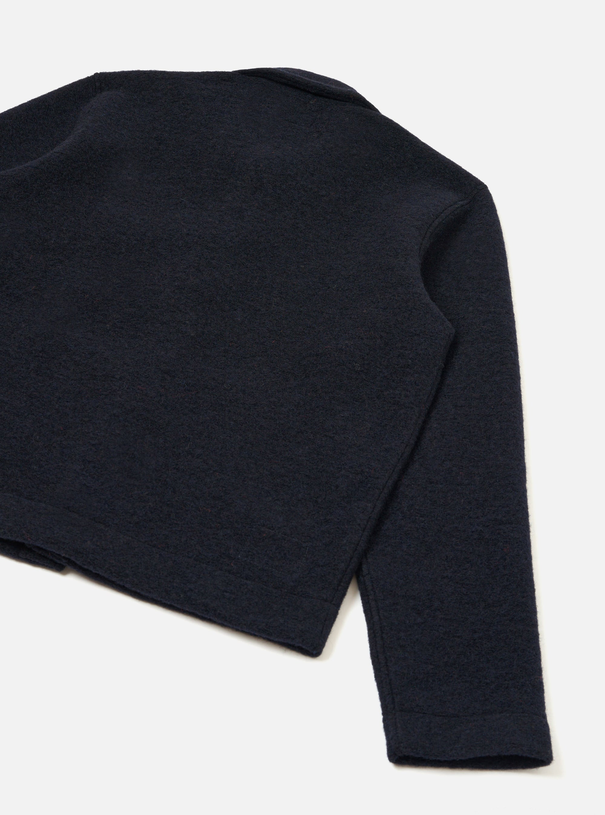 Flat lay of a navy wool fleece uniform jacket shot from above at an angle on a white surface. The back and one sleeve are visible, showing the boiled wool texture, a clean ribbed hem, and a small collar visible at the top. The sleeve tapers to a plain cuff.