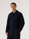 Navy wool fleece uniform jacket worn buttoned by a male model, cropped at mid-thigh. Features a spread collar, five dark buttons down the front placket, a chest patch pocket, and two lower patch pockets. Textured fleece fabric throughout. Worn over a grey crew-neck tee with navy trousers. Grey studio backdrop.