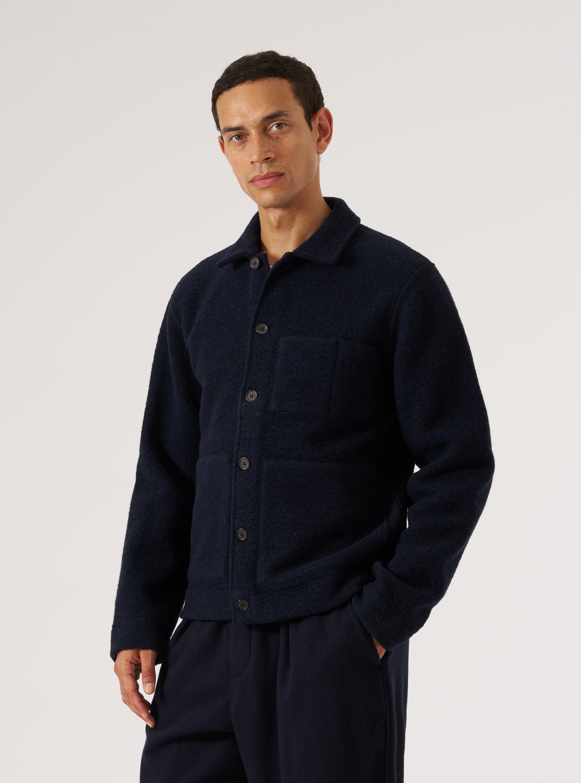 Navy wool fleece uniform jacket worn buttoned by a male model, cropped at mid-thigh. Features a spread collar, five dark buttons down the front placket, a chest patch pocket, and two lower patch pockets. Textured fleece fabric throughout. Worn over a grey crew-neck tee with navy trousers. Grey studio backdrop.