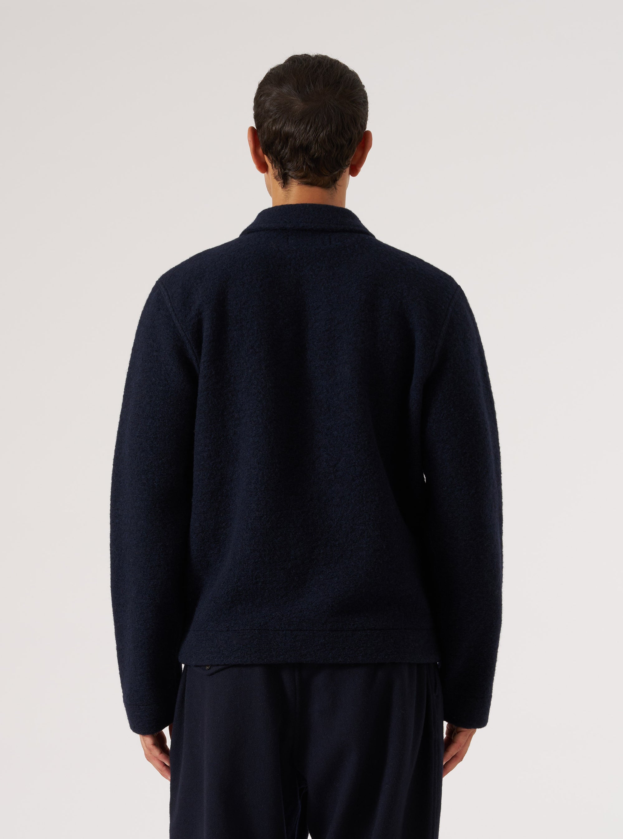 Back view of a male model wearing a navy wool fleece uniform jacket against a light grey studio backdrop. The textured fleece fabric is visible across the back and full-length sleeves. A spread collar is visible at the neckline. The jacket sits at hip length with a ribbed hem. Worn over navy trousers. Shot cropped above the knees.