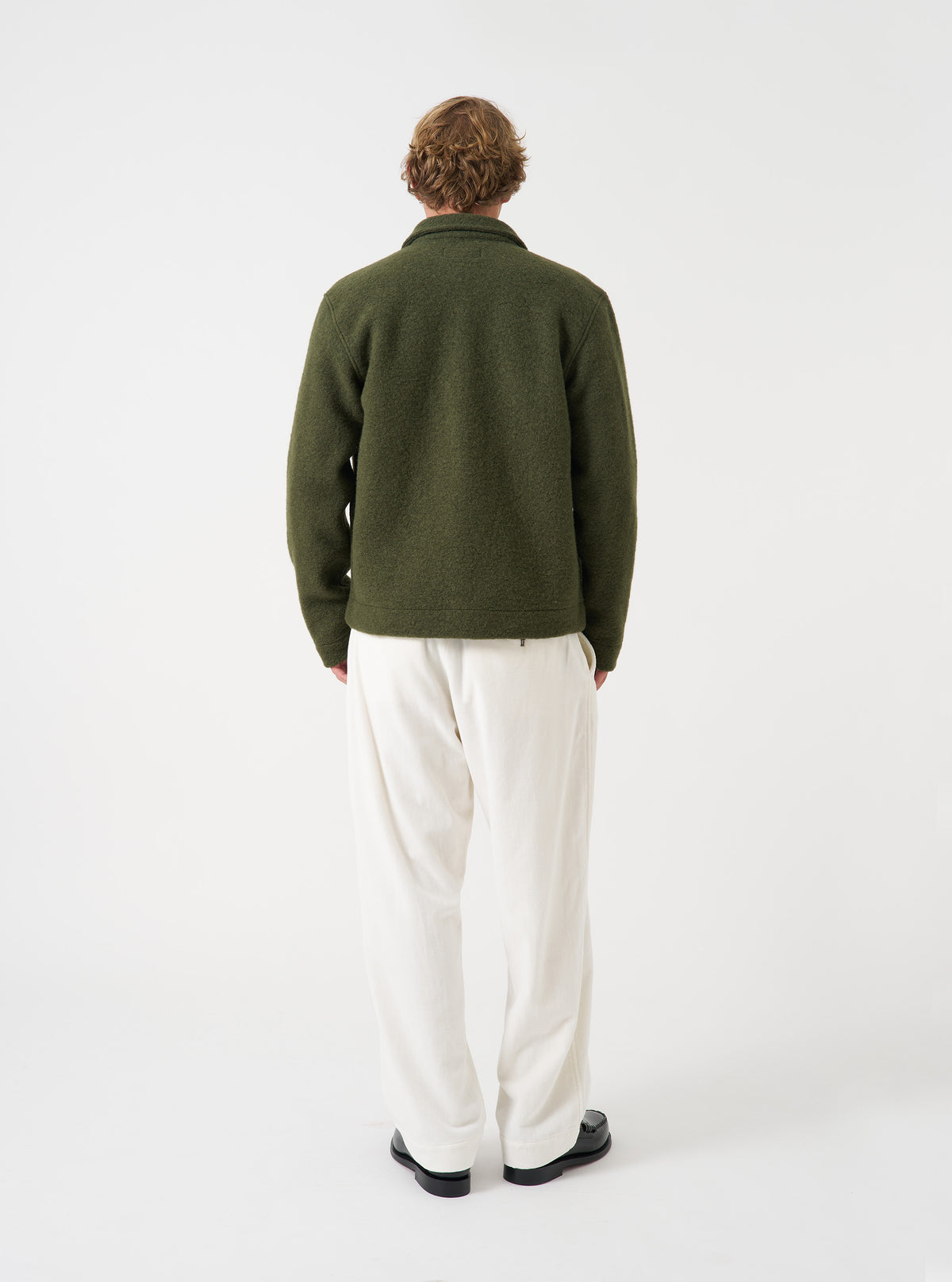 Back view of a model wearing the Olive Wool Fleece Uniform Jacket on a plain studio backdrop. The shot showcases the straight cut hemline, smooth back panel construction, and green olive wool fleece fabric structure.