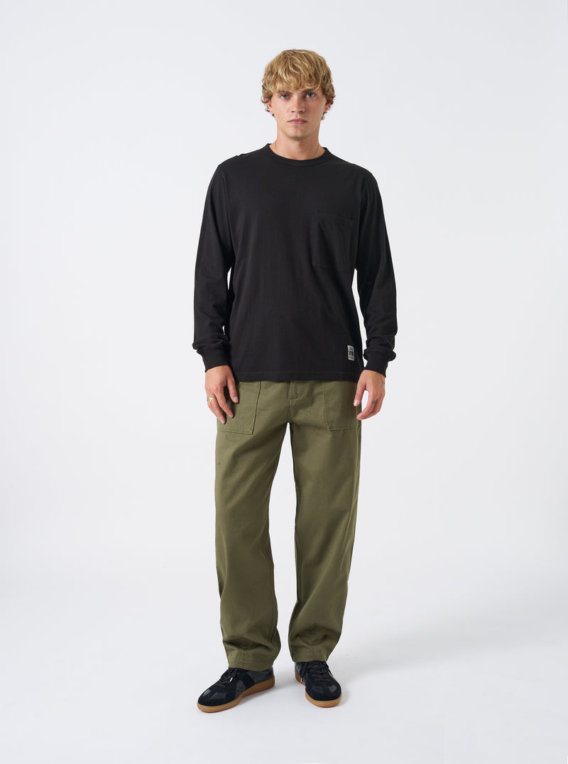 Front view of a model wearing the Black Single Jersey L/S Core Pocket Tee against a plain light studio backdrop. The model faces front, displaying the black cotton jersey shirt, long sleeves, crew neck, and chest pocket.