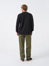 Back view of a model wearing the Black Single Jersey L/S Core Pocket Tee against a plain light studio backdrop. The image focuses on the reverse side of the black long-sleeve T-shirt, showing shoulder seams and clean hem.