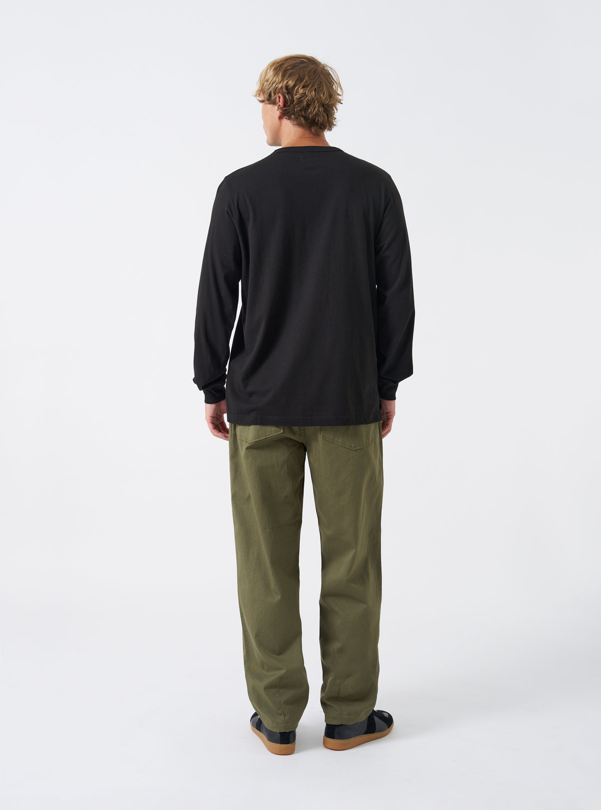 Back view of a model wearing the Black Single Jersey L/S Core Pocket Tee against a plain light studio backdrop. The image focuses on the reverse side of the black long-sleeve T-shirt, showing shoulder seams and clean hem.