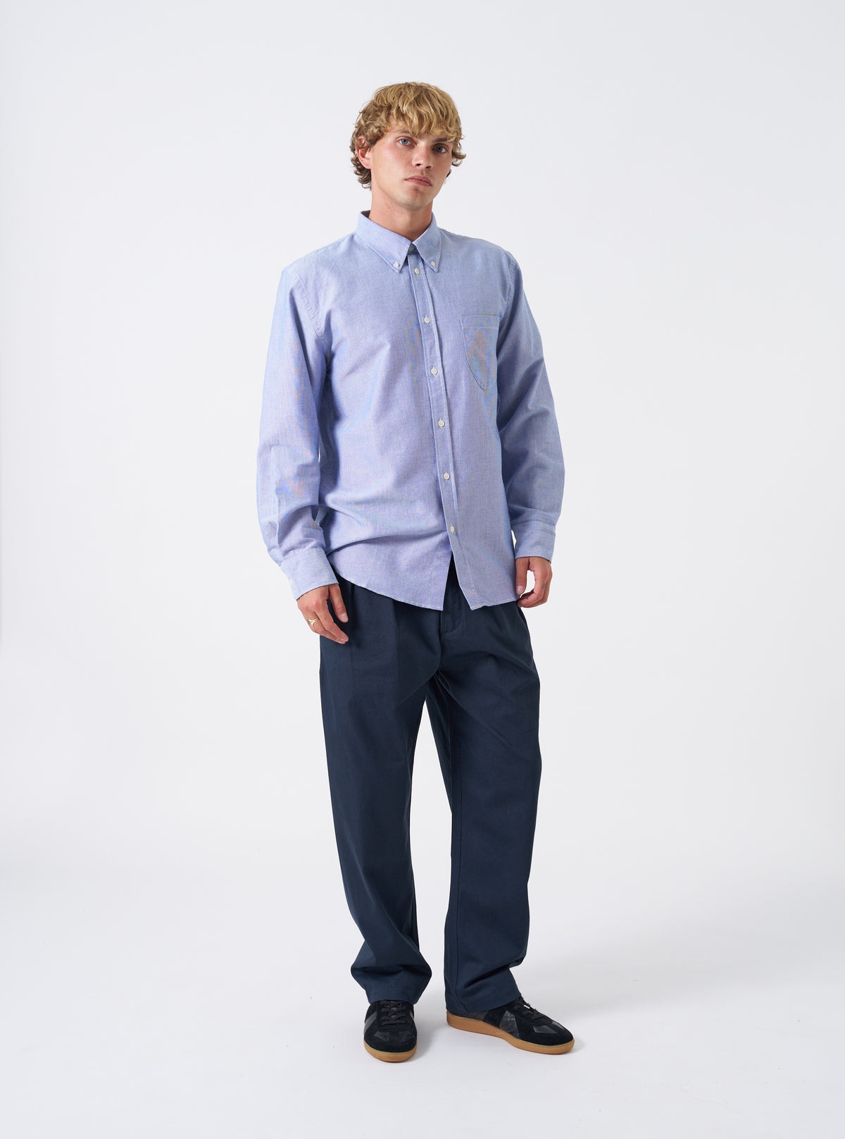 Front view of a model wearing the Blue Oxford Daybrook Shirt paired with trousers in a studio setting. The model faces forward, showcasing how the blue organic oxford button-down shirt fits across the chest and shoulders.