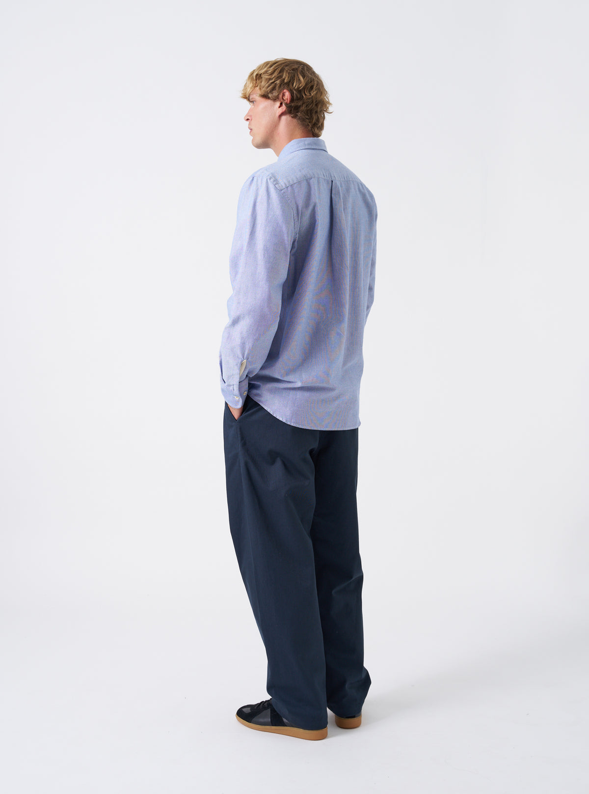 Side angled view of a model wearing the Blue Oxford Daybrook Shirt against a neutral backdrop. The shot illustrates the long-sleeve cuff detail, relaxed tailored silhouette, and collar structure from a side angle.