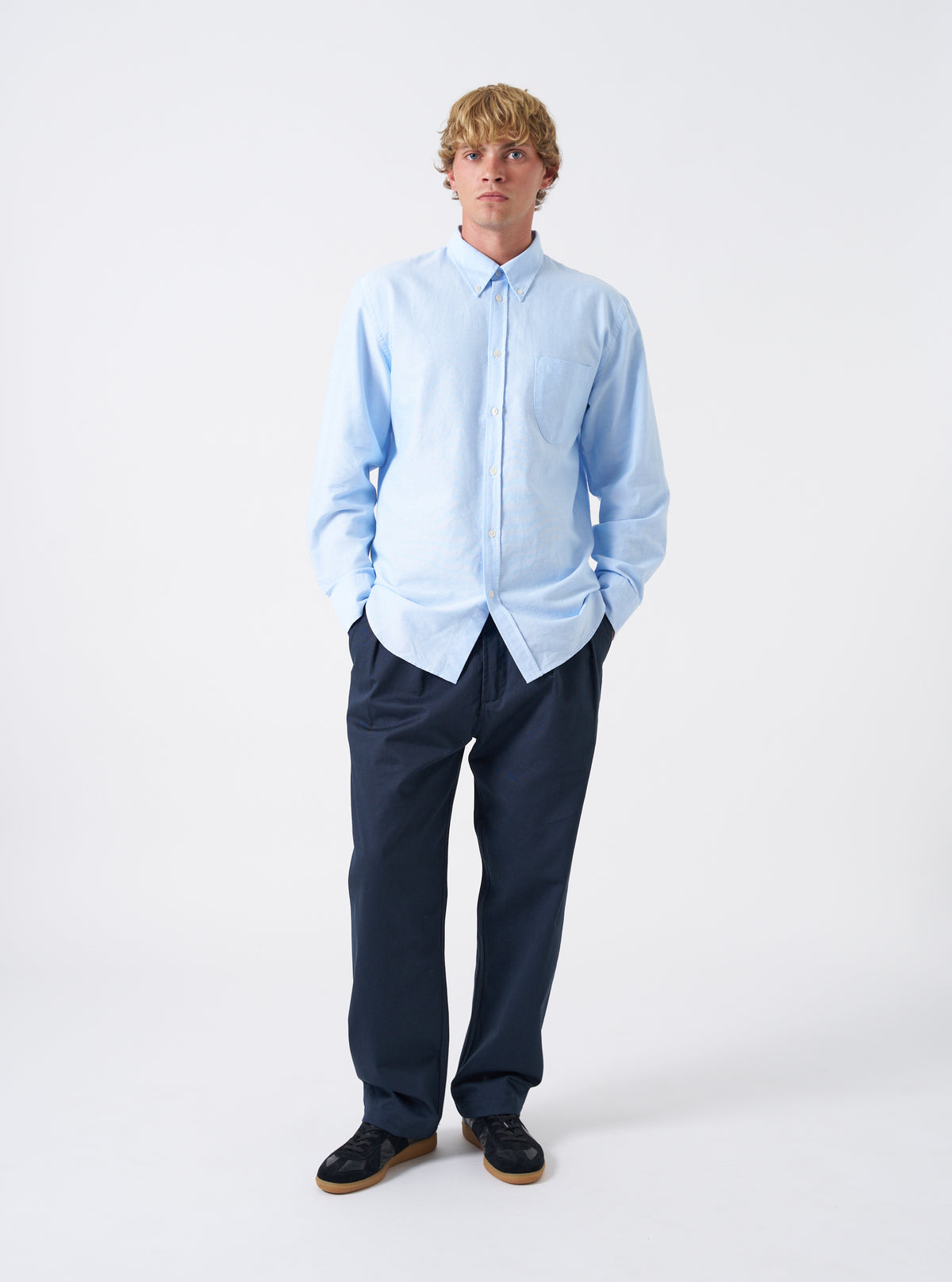 Front view of a model wearing the Sky Blue Oxford Daybrook Shirt in a minimalist studio setting. The model stands facing the camera, showing the overall fit, collar position, and light blue tone of the long-sleeve shirt.