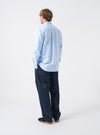 Angled view of a model wearing the Sky Blue Oxford Daybrook Shirt against a plain studio background. Highlights the side profile fit, sleeve drape, cuff finish, and classic casual look of the organic oxford garment.