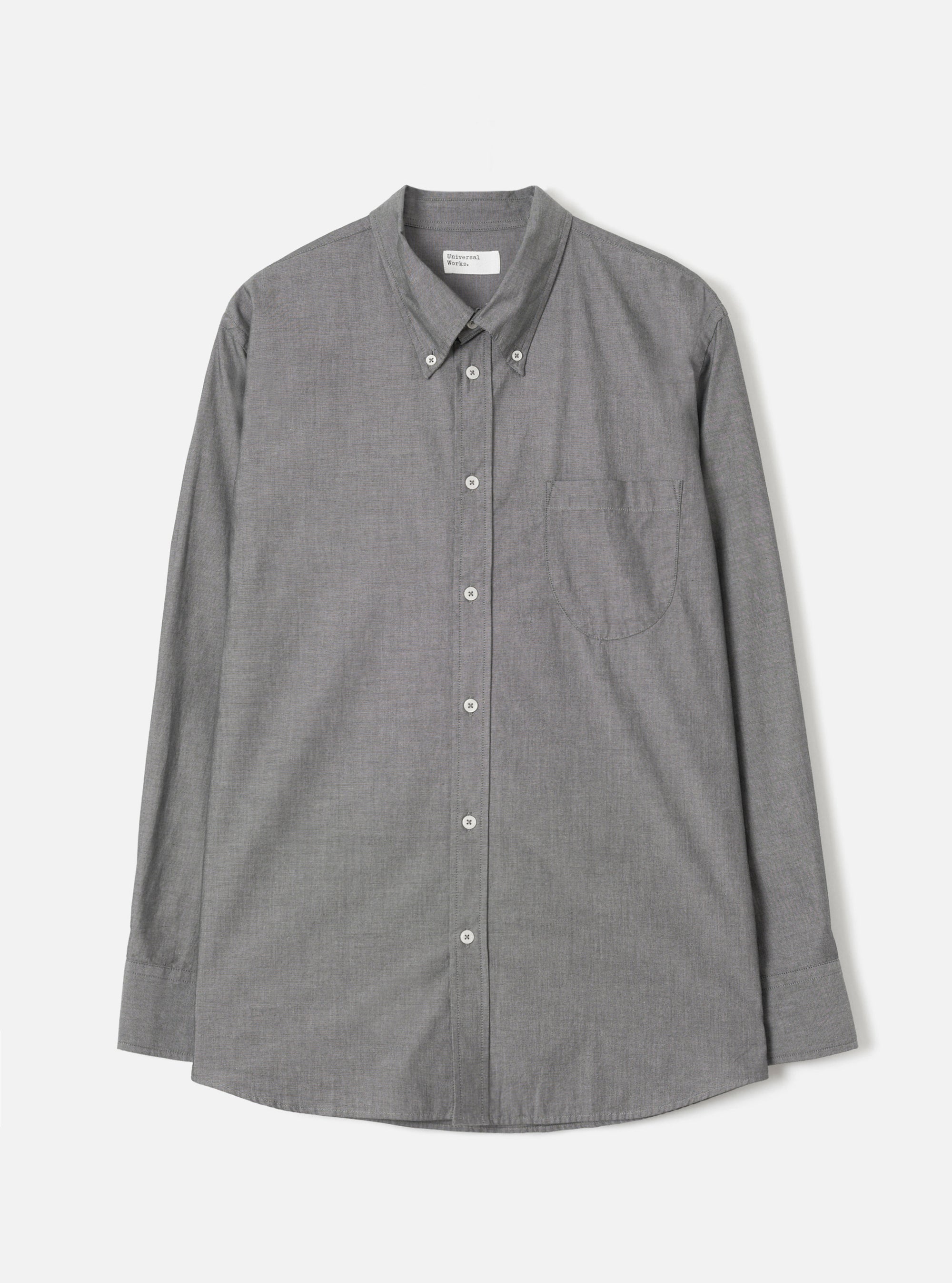 Grey chambray Daybrook shirt by Universal Works, flat lay on a light grey seamless backdrop. Button-down collar with buttoned collar points, full button placket with white buttons, single chest patch pocket, and long sleeves with buttoned cuffs. Fine woven texture visible throughout. Curved hem. Universal Works woven label at neck.
