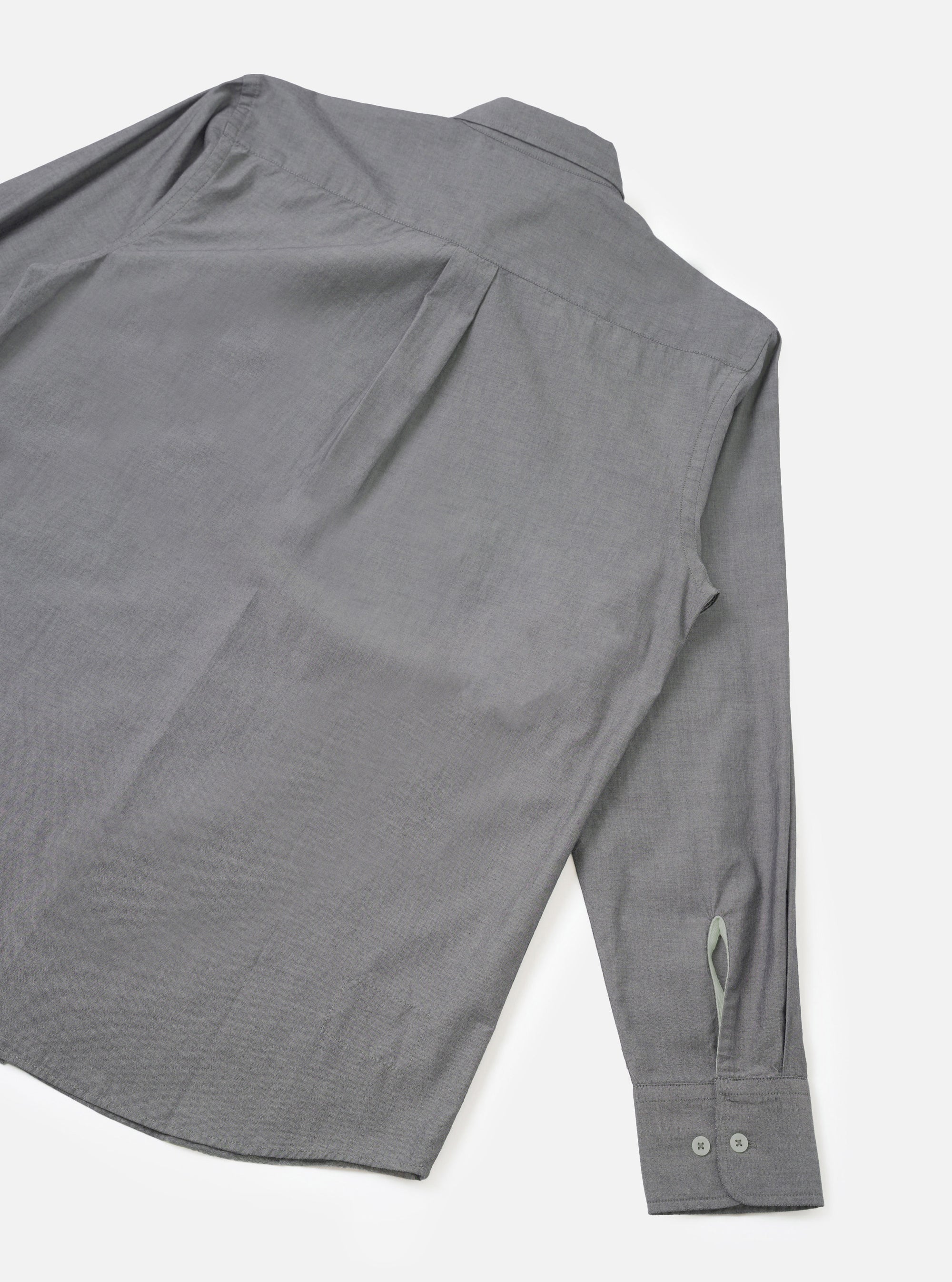 Flat lay of the Black Prince chambray Daybrook shirt on a white studio backdrop, showing the back and one sleeve. The fine-weave grey chambray fabric has a subtle texture. A back yoke seam runs across the shoulders. The cuff is buttoned with two small grey buttons and features a placket opening.