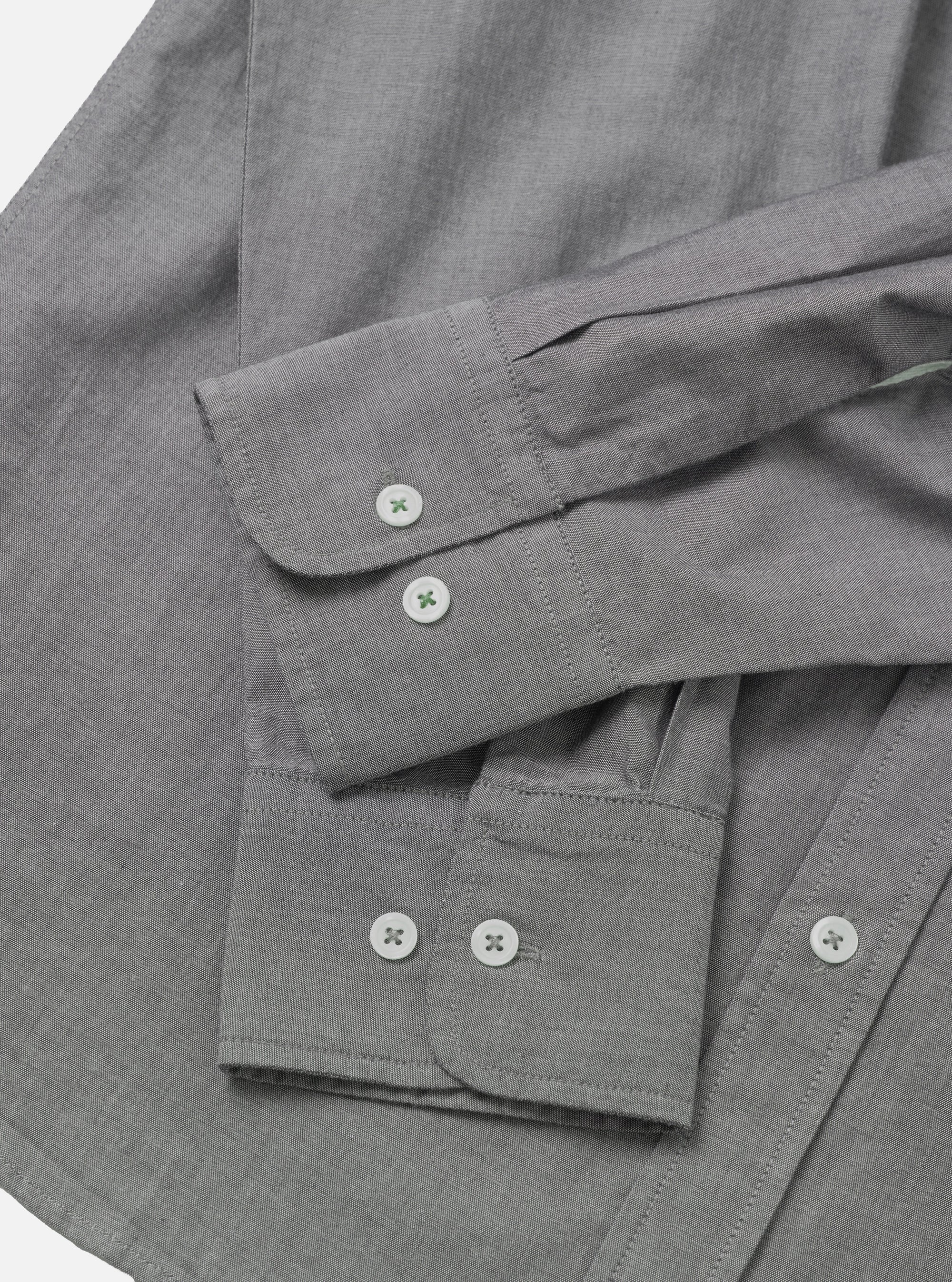 Close-up flat lay of a grey chambray shirt on a white surface, showing two barrel cuffs with white buttons stitched in green thread. The foreground cuff has two buttons side by side; the background cuff has one visible button. Top-stitching runs along the cuff edges. The fine chambray weave texture is visible across the body of the shirt.