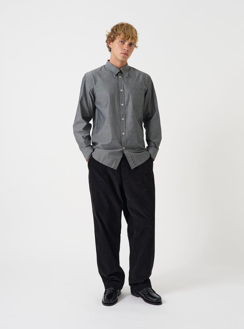 A male model poses wearing the Black Prince Chambray Daybrook Shirt buttoned up against a plain light grey backdrop. The long-sleeve cotton shirt features a pointed collar and chest pocket, styled with dark wide-leg trousers.