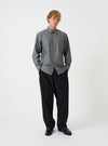 A male model poses wearing the Black Prince Chambray Daybrook Shirt buttoned up against a plain light grey backdrop. The long-sleeve cotton shirt features a pointed collar and chest pocket, styled with dark wide-leg trousers.