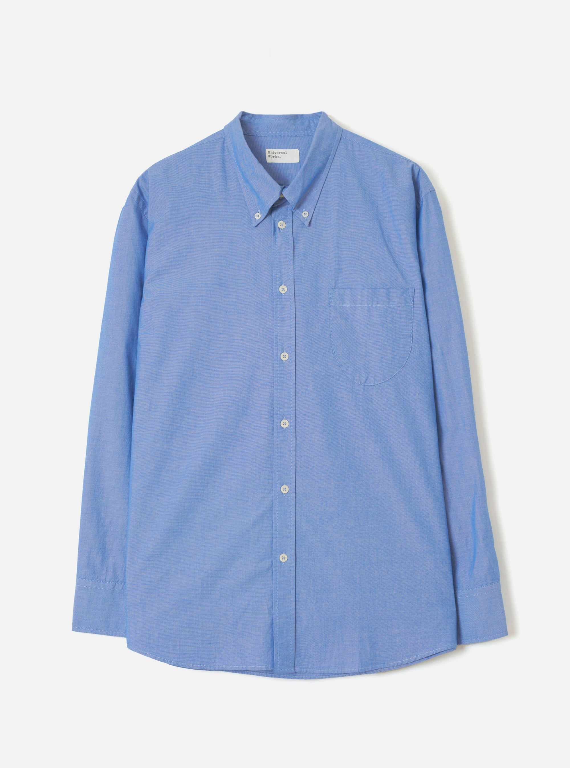 Blue chambray shirt laid flat on a light grey backdrop. Features a button-down collar, front button placket with white buttons, a single chest patch pocket, long sleeves with buttoned cuffs, and a curved hem. The fine woven texture of the chambray fabric is visible throughout.