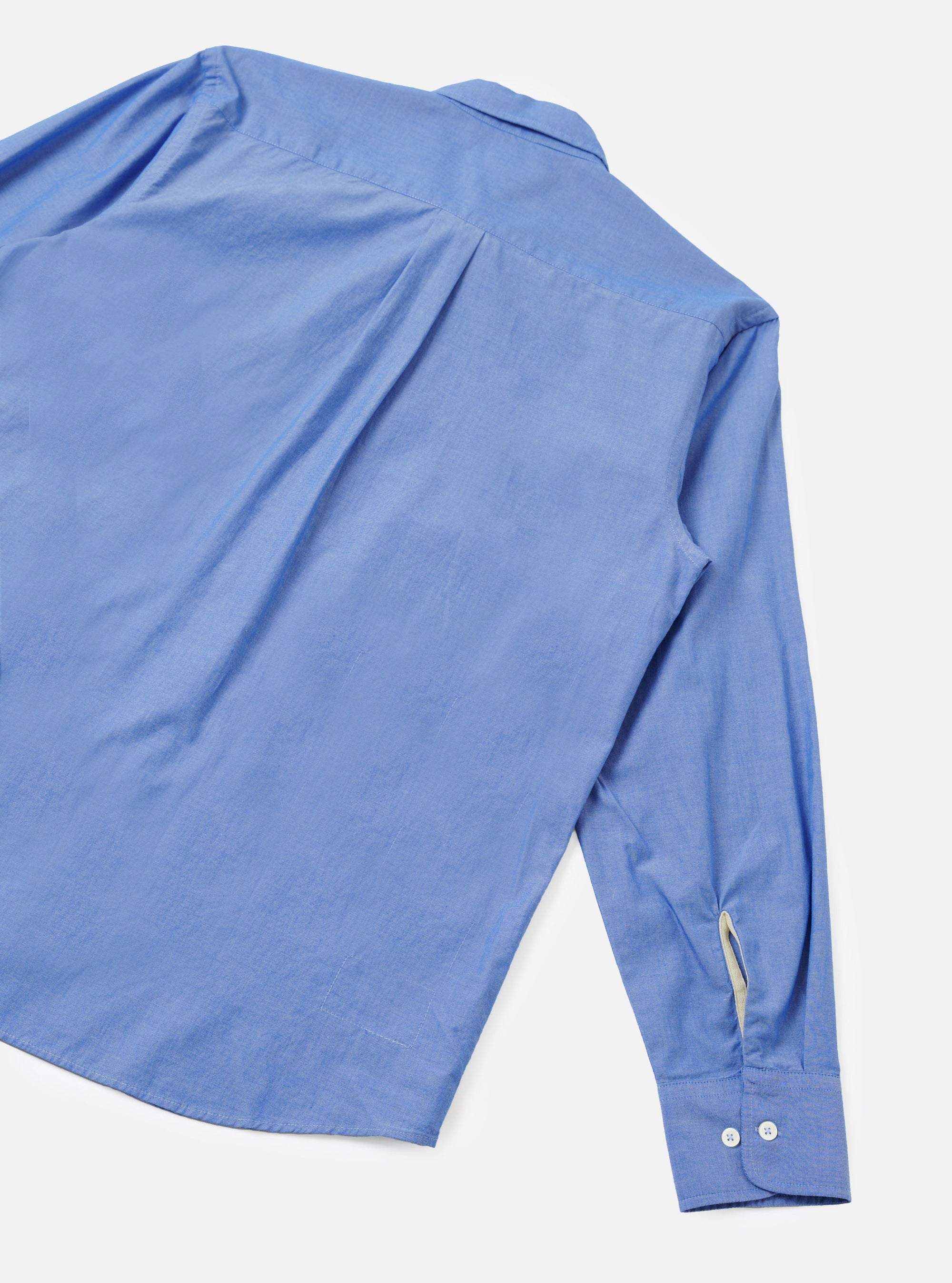 Blue chambray Daybrook shirt laid flat on a light grey seamless backdrop, shot from above at an angle. The back yoke, point collar and one long sleeve are visible. The buttoned cuff shows two white buttons, with a cream contrast placket detail at the sleeve opening.