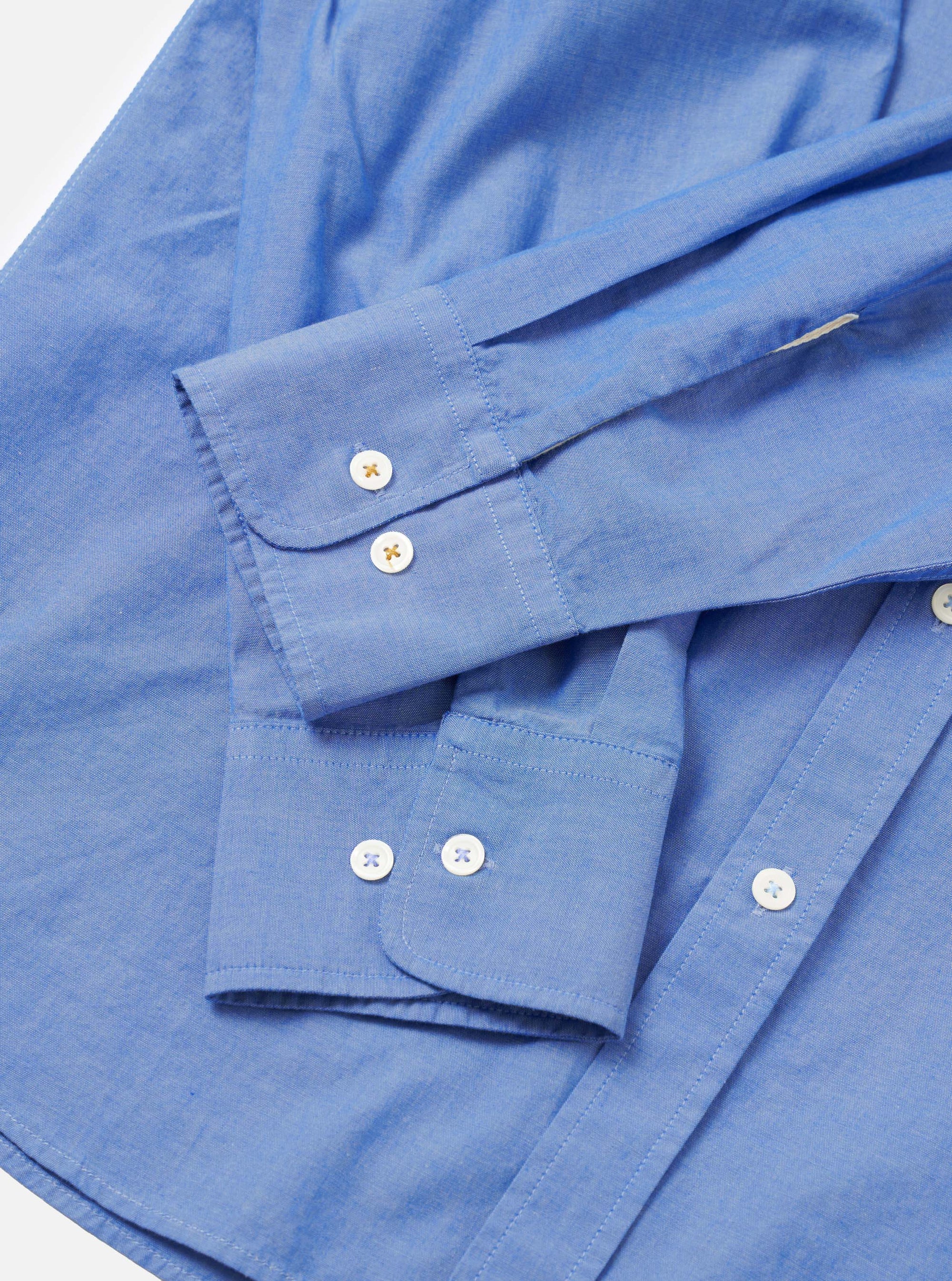 Close-up flat lay of the blue chambray Daybrook shirt on a white surface, focusing on two long sleeves and their cuffs. Each barrel cuff features two cream buttons with cross stitching. Top-stitching details the cuff edges and placket. The fine chambray weave is visible throughout.