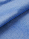 Close-up of the blue chambray fabric used in the Daybrook shirt, folded diagonally to show texture and drape. The plain-weave oxford-style cloth shows a fine two-tone weave of blue and white threads, giving a soft, mottled finish. Shot against a white studio backdrop.