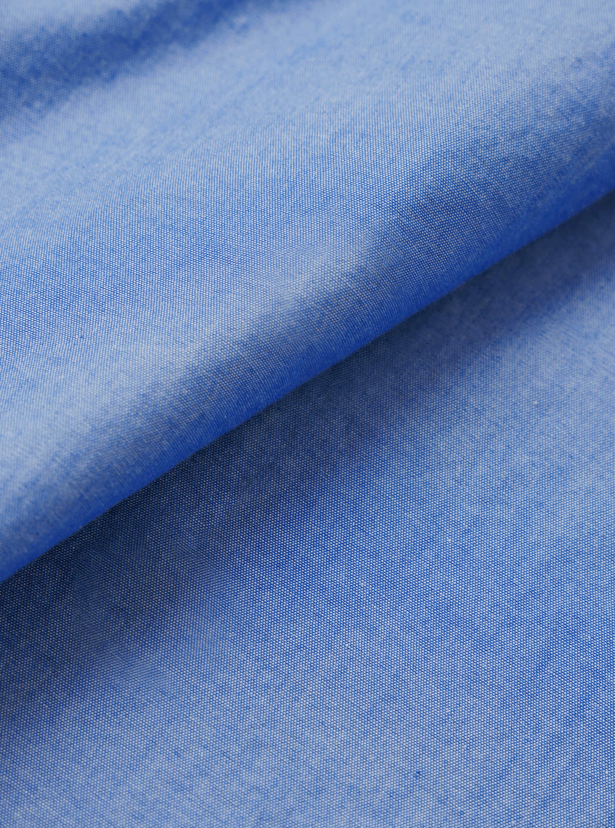 Close-up of the blue chambray fabric used in the Daybrook shirt, folded diagonally to show texture and drape. The plain-weave oxford-style cloth shows a fine two-tone weave of blue and white threads, giving a soft, mottled finish. Shot against a white studio backdrop.