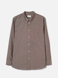 Brown chambray long-sleeve shirt laid flat on a light grey studio backdrop. Button-down collar with a Universal Works woven label at the back neck. Seven pale buttons down the placket, one chest patch pocket on the left, and single-button barrel cuffs. Fine woven texture with a subtle two-tone weave. Curved hem.