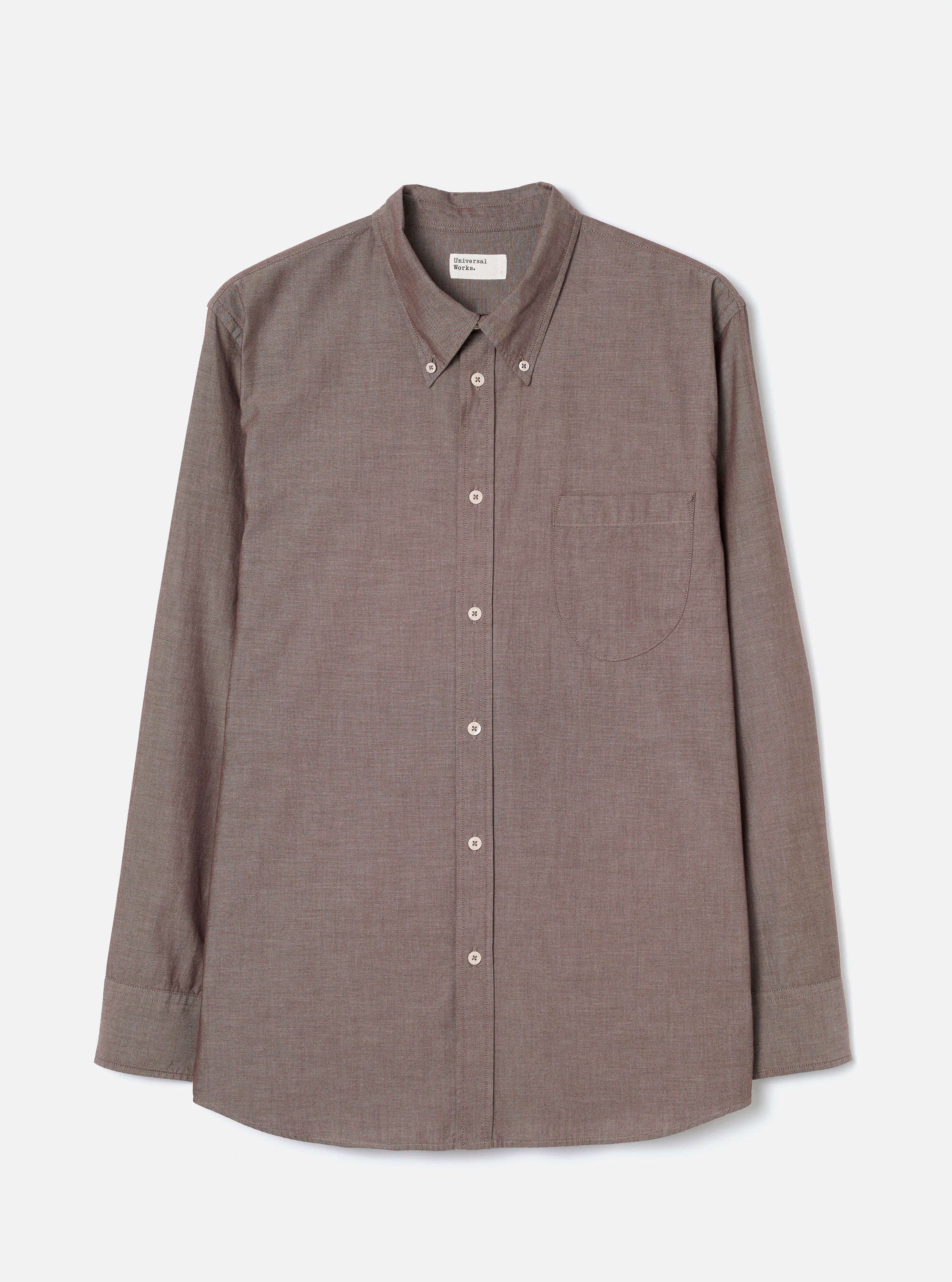 Brown chambray long-sleeve shirt laid flat on a light grey studio backdrop. Button-down collar with a Universal Works woven label at the back neck. Seven pale buttons down the placket, one chest patch pocket on the left, and single-button barrel cuffs. Fine woven texture with a subtle two-tone weave. Curved hem.