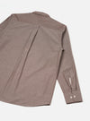 Flat lay of the brown chambray Daybrook shirt showing the back. A centre back pleat runs from the yoke to the curved hem. One long sleeve is visible, with a buttoned barrel cuff fastened with two cream buttons. The collar is partially visible at the top. Fine chambray weave texture is visible throughout. White studio backdrop.