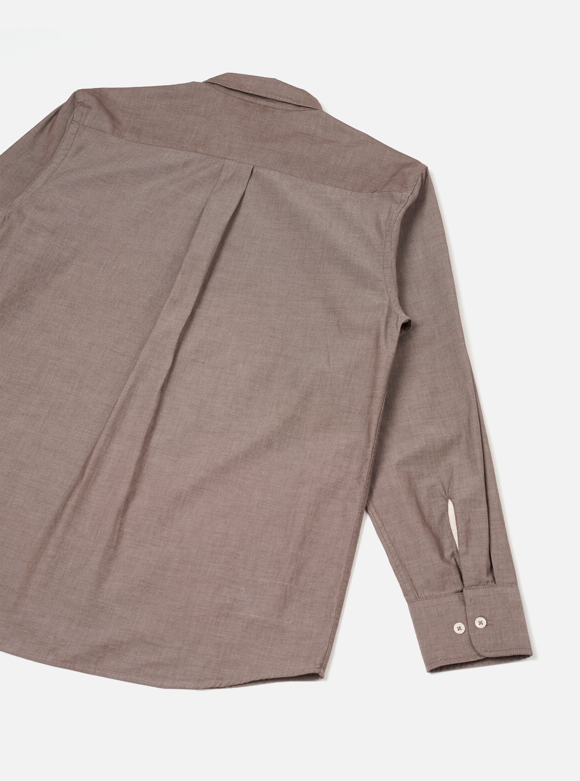 Flat lay of the brown chambray Daybrook shirt showing the back. A centre back pleat runs from the yoke to the curved hem. One long sleeve is visible, with a buttoned barrel cuff fastened with two cream buttons. The collar is partially visible at the top. Fine chambray weave texture is visible throughout. White studio backdrop.