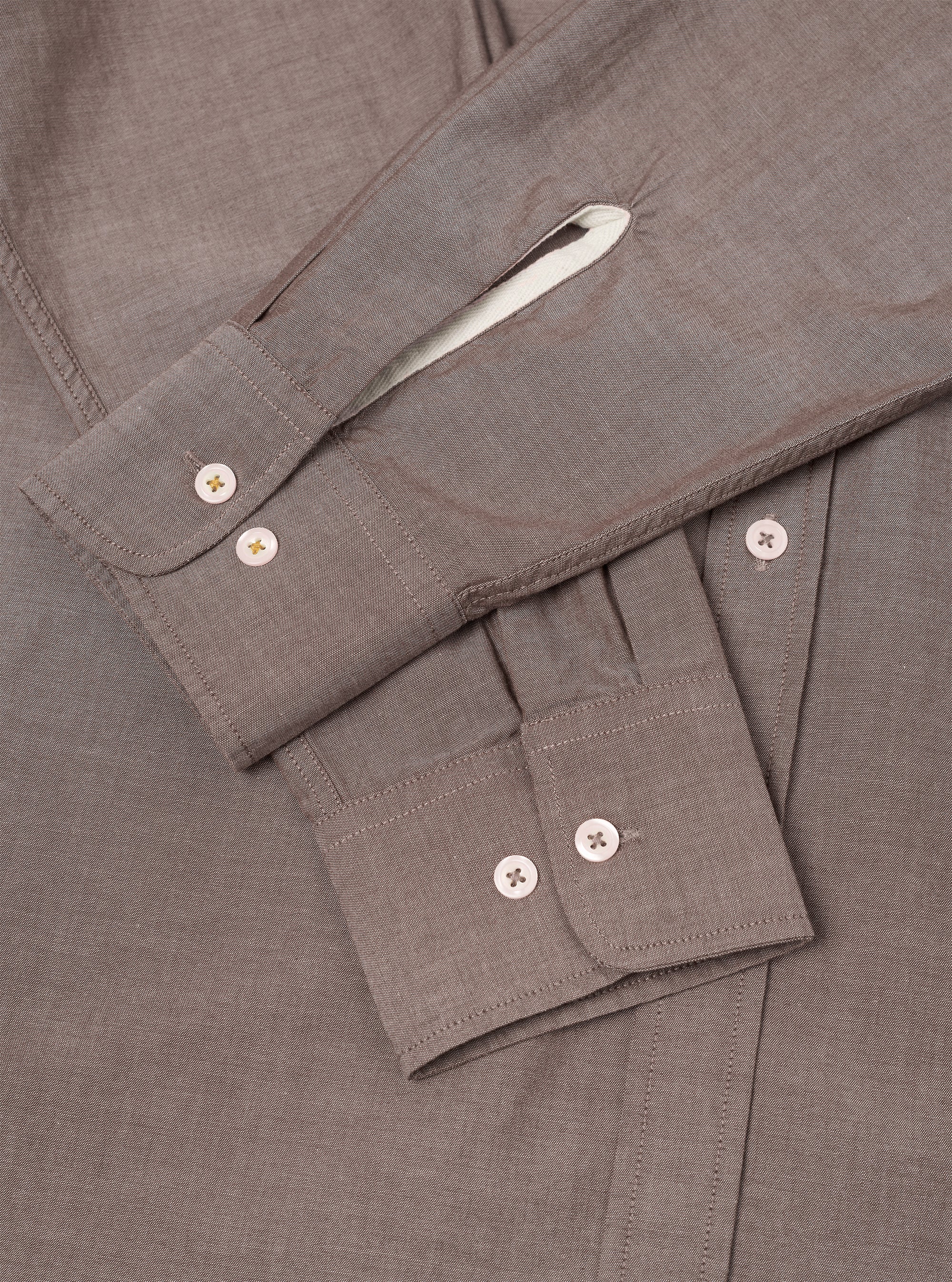 Close-up flat lay of a brown chambray shirt, focusing on the sleeve and cuff detail. Two barrel cuffs each fasten with two cream buttons. Topstitching outlines the cuffs and placket. A white contrast lining is visible inside the folded collar. The fine chambray weave is visible across the fabric.