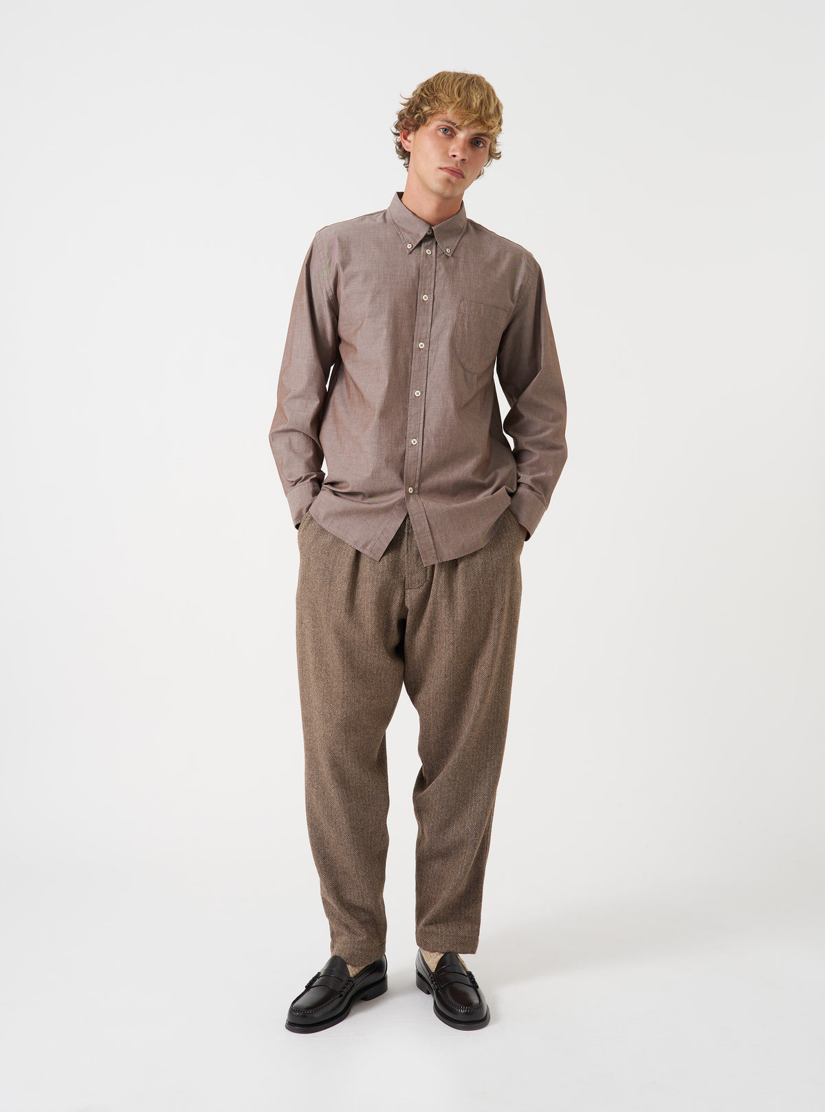 A male model poses against a plain studio backdrop wearing the Brown Prince Chambray Daybrook Shirt. The long-sleeve button-up shirt features a neat collar, a single chest pocket, and a clean front closure.