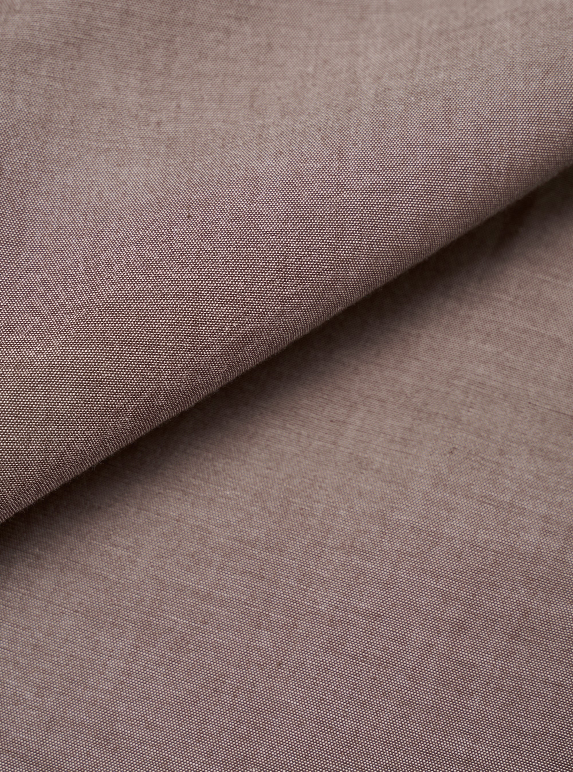 Close-up of the brown chambray fabric used in the Daybrook Shirt. The plain-weave textile shows a fine two-tone woven texture with subtle natural slubs across the surface. The fabric is folded diagonally, revealing depth and drape. No garment construction details are visible.