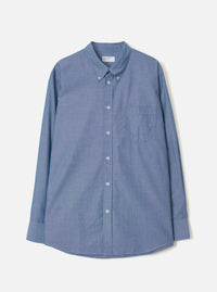 Indigo chambray long-sleeve shirt laid flat on a light grey background. Features a button-down collar, centre-front button placket with white buttons, a left-chest patch pocket, and barrel cuffs. The curved shirttail hem is visible at the base. Fine chambray weave texture throughout.