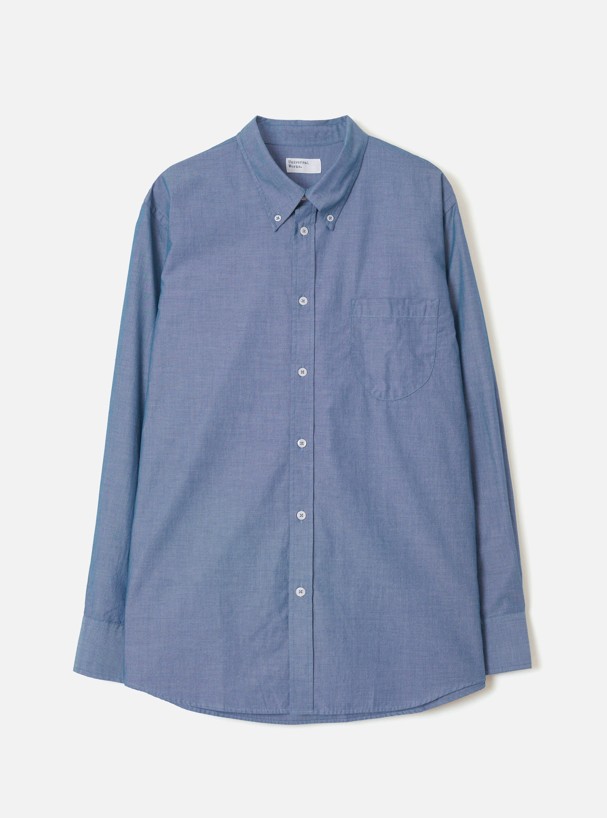 Indigo chambray long-sleeve shirt laid flat on a light grey background. Features a button-down collar, centre-front button placket with white buttons, a left-chest patch pocket, and barrel cuffs. The curved shirttail hem is visible at the base. Fine chambray weave texture throughout.