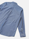 Rear flat-lay of the indigo chambray Daybrook shirt on a light grey seamless backdrop. The fine woven chambray fabric shows a subtle two-tone texture. Visible details include a back box pleat below the collar, a spread collar, a long sleeve with a neat barrel cuff fastened with two small grey buttons.