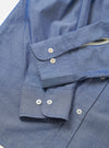 Close-up flat lay of the indigo chambray Daybrook shirt on a white surface, focusing on both cuffs. Each rounded cuff fastens with two mother-of-pearl buttons with coloured cross-stitching. The placket on the right shows two further buttons. Fine chambray weave texture is visible throughout, with a cream contrast lining visible inside one cuff.