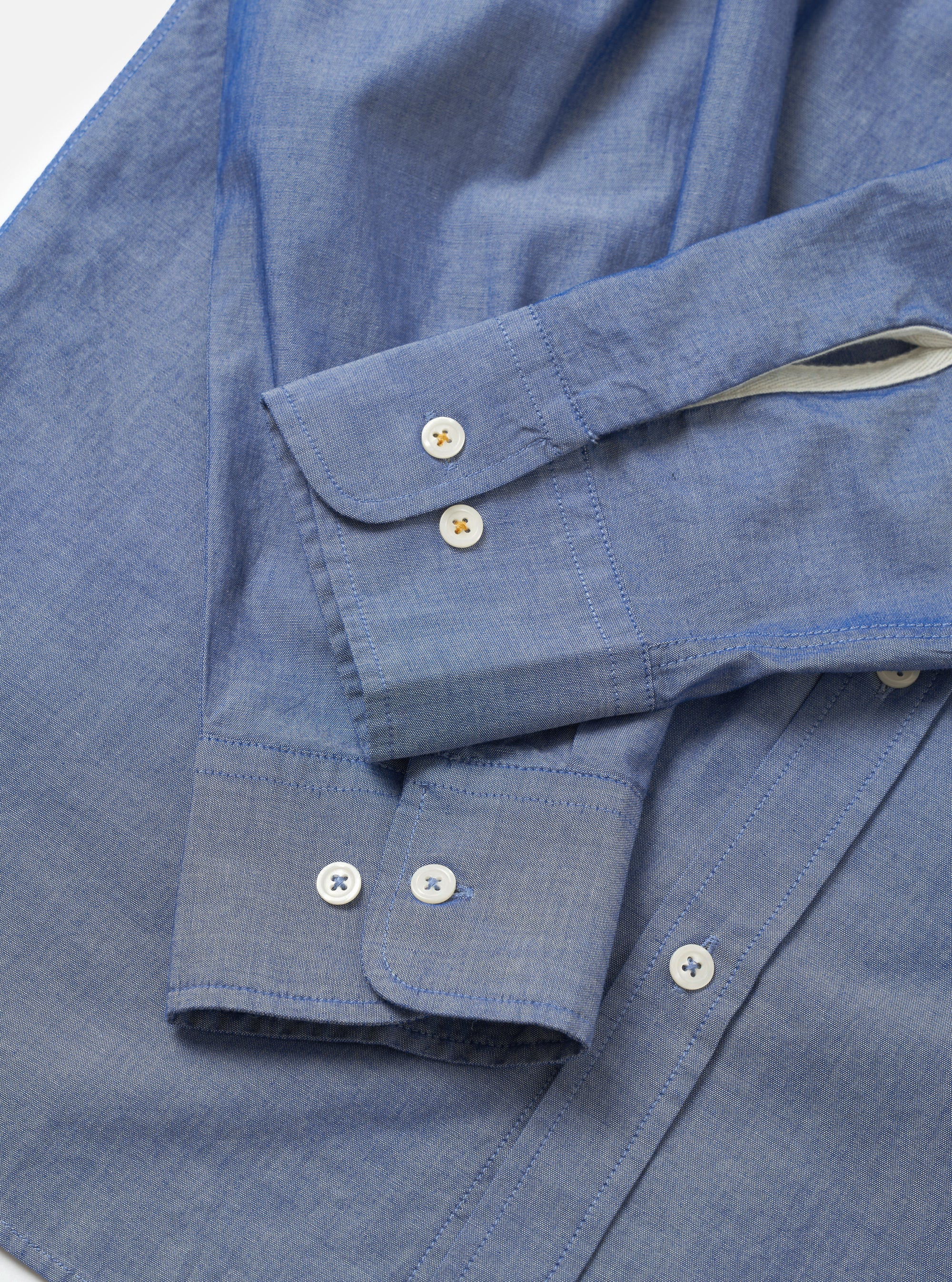 Close-up flat lay of the indigo chambray Daybrook shirt on a white surface, focusing on both cuffs. Each rounded cuff fastens with two mother-of-pearl buttons with coloured cross-stitching. The placket on the right shows two further buttons. Fine chambray weave texture is visible throughout, with a cream contrast lining visible inside one cuff.