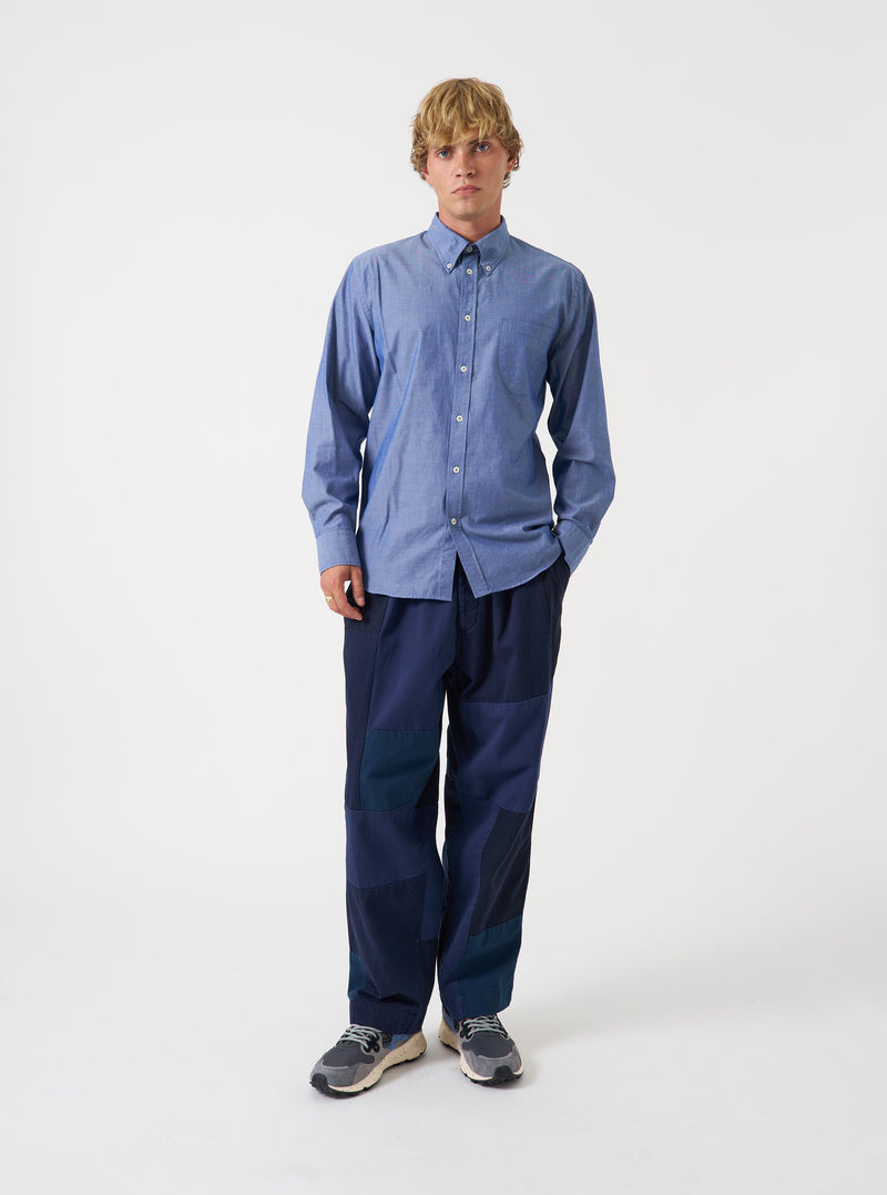A male model poses against a light grey studio backdrop wearing the Indigo Prince Chambray Daybrook Shirt. The classic long-sleeve button-down shirt features a point collar and front patch pocket, worn unbuttoned over a plain white tee.