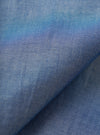 Close-up of the indigo chambray fabric used in the Daybrook shirt, showing a fine two-tone weave of blue and white threads. A diagonal fold runs across the frame, highlighting the fabric's soft drape and subtle texture variation across the surface.
