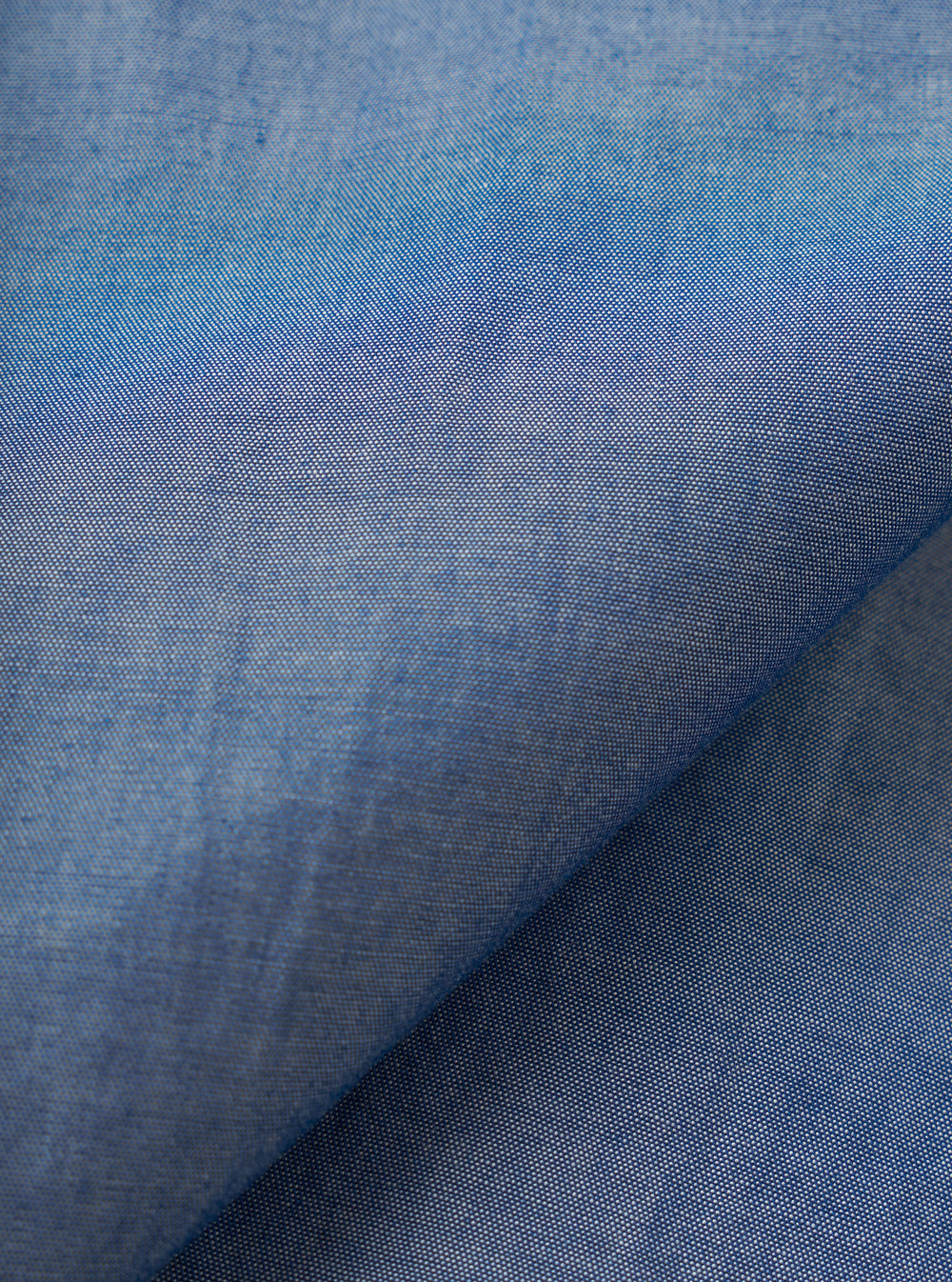Close-up of the indigo chambray fabric used in the Daybrook shirt, showing a fine two-tone weave of blue and white threads. A diagonal fold runs across the frame, highlighting the fabric's soft drape and subtle texture variation across the surface.