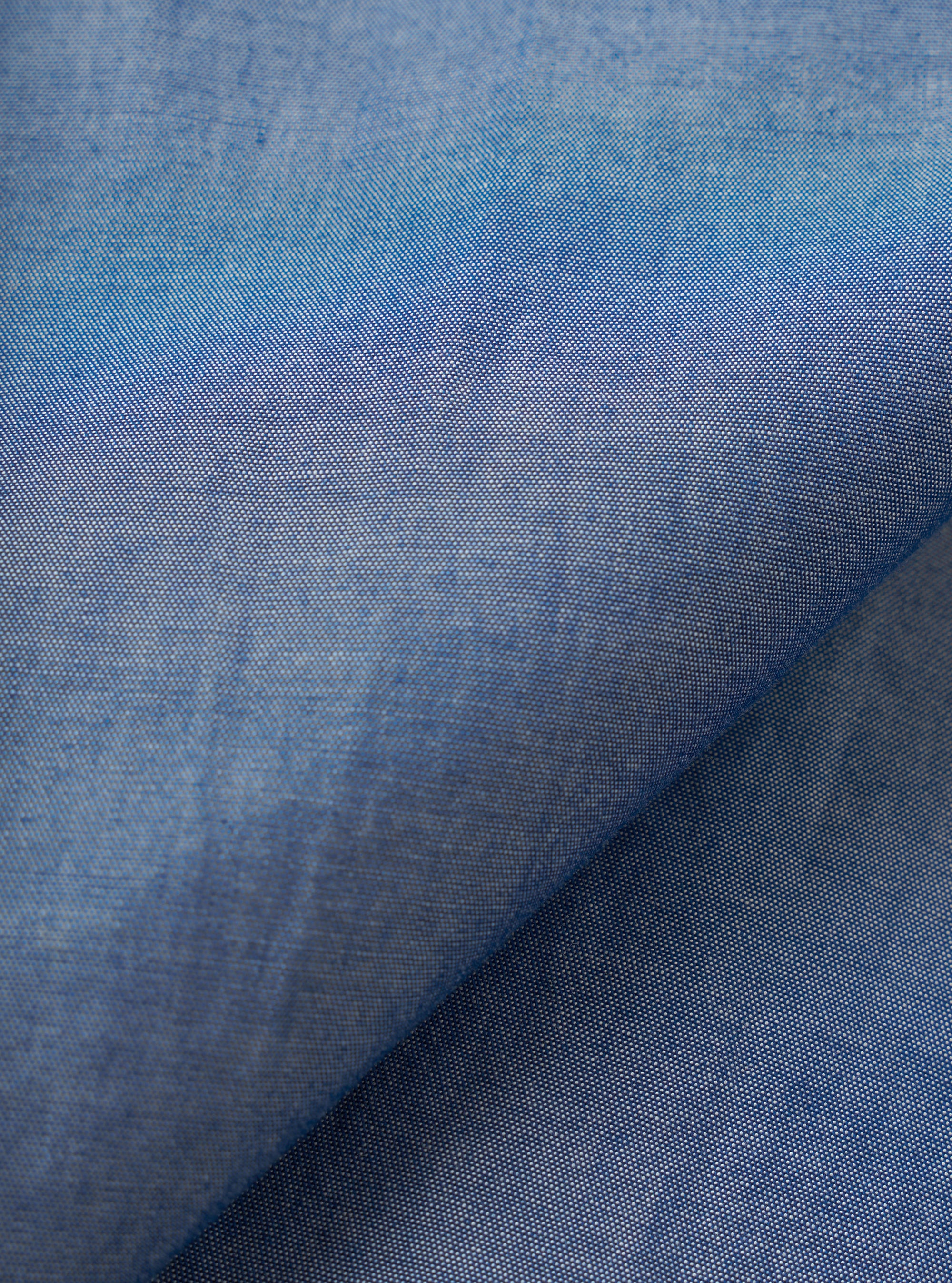 Close-up of the indigo chambray fabric used in the Daybrook shirt, showing a fine two-tone weave of blue and white threads. A diagonal fold runs across the frame, highlighting the fabric's soft drape and subtle texture variation across the surface.