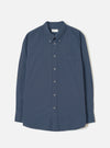 Navy chambray long-sleeve shirt laid flat on a light grey studio backdrop. Features a button-down collar, buttoned placket with six grey buttons, a curved rounded hem, single chest patch pocket, and single-button barrel cuffs. A branded woven label sits at the centre back neck.