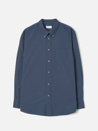 Navy chambray long-sleeve shirt laid flat on a light grey studio backdrop. Features a button-down collar, buttoned placket with six grey buttons, a curved rounded hem, single chest patch pocket, and single-button barrel cuffs. A branded woven label sits at the centre back neck.