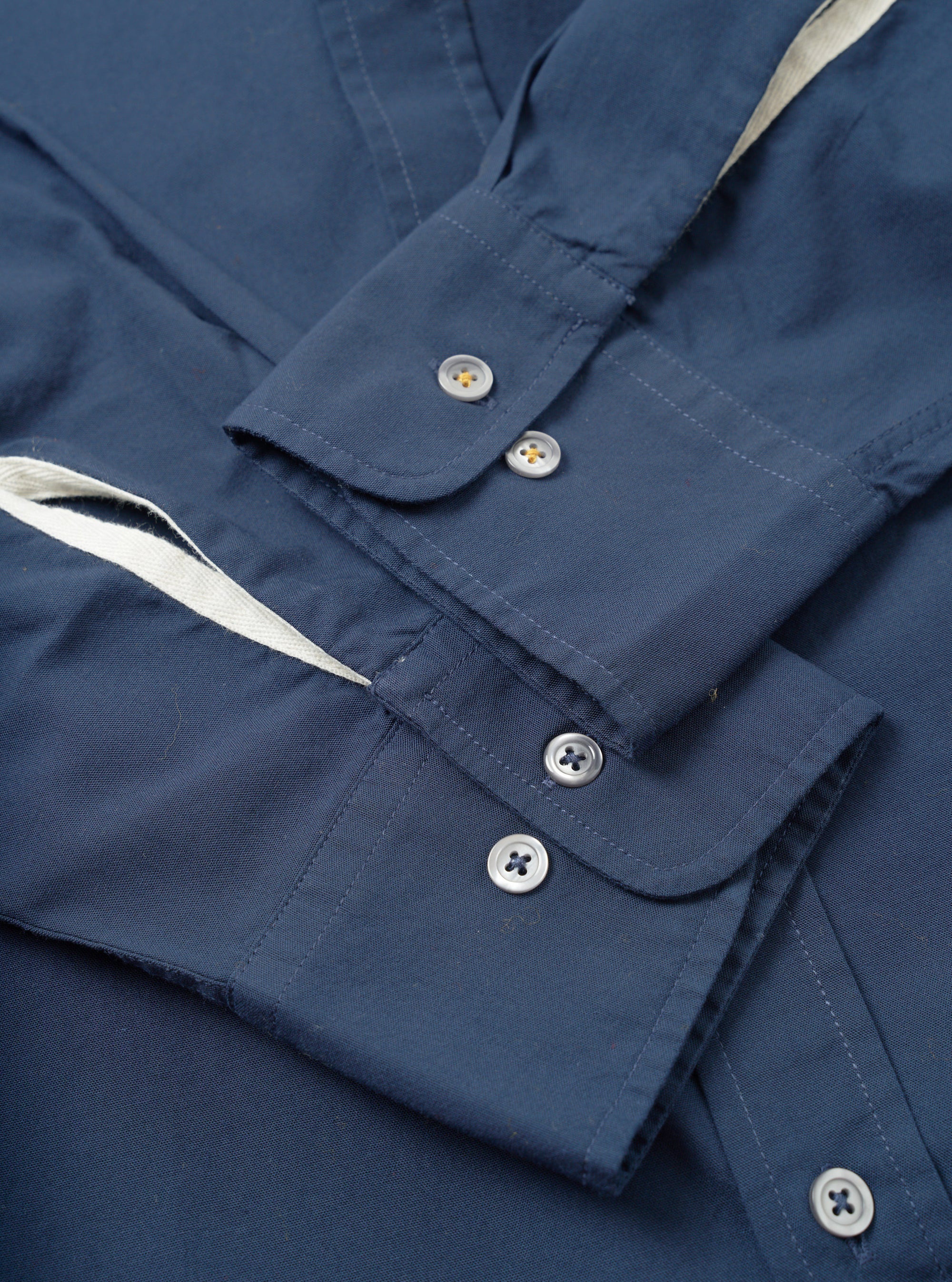 Close-up flat lay of the navy chambray Daybrook shirt showing two buttoned barrel cuffs with silver four-hole buttons. A cream cotton twill tape trim is visible along the placket edge. The plain-weave fabric has a fine texture. Top-stitching details the cuff edges. Shot on a white studio surface.