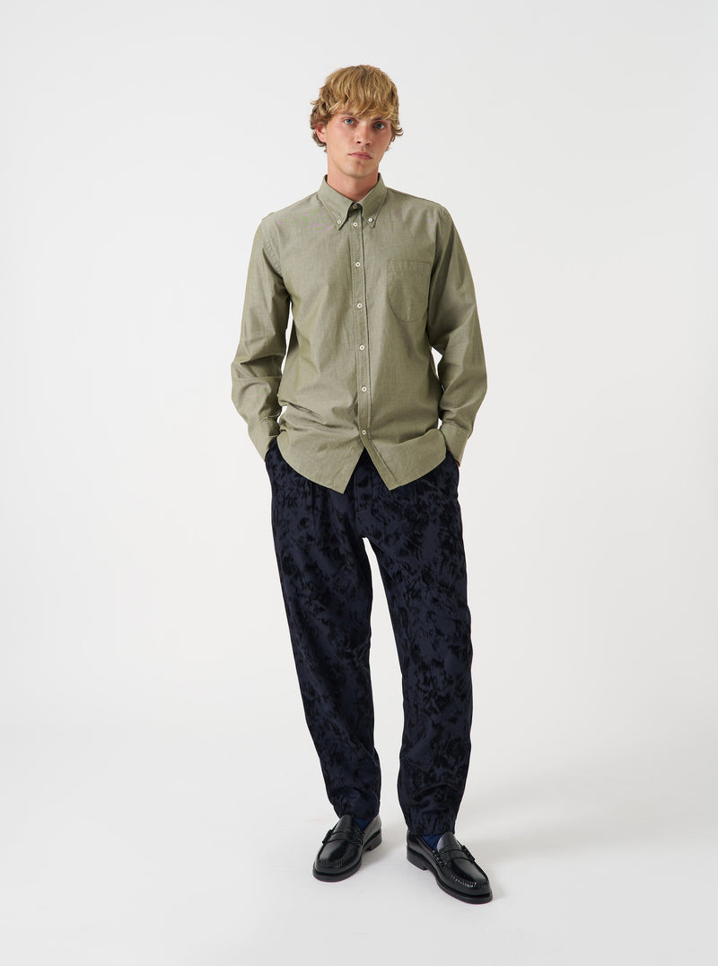 A male model poses against a plain studio backdrop wearing the Olive Prince Chambray Daybrook Shirt. The shirt features an olive green cotton weave, a spread collar, button-up front closure, and a single patch pocket at the chest.