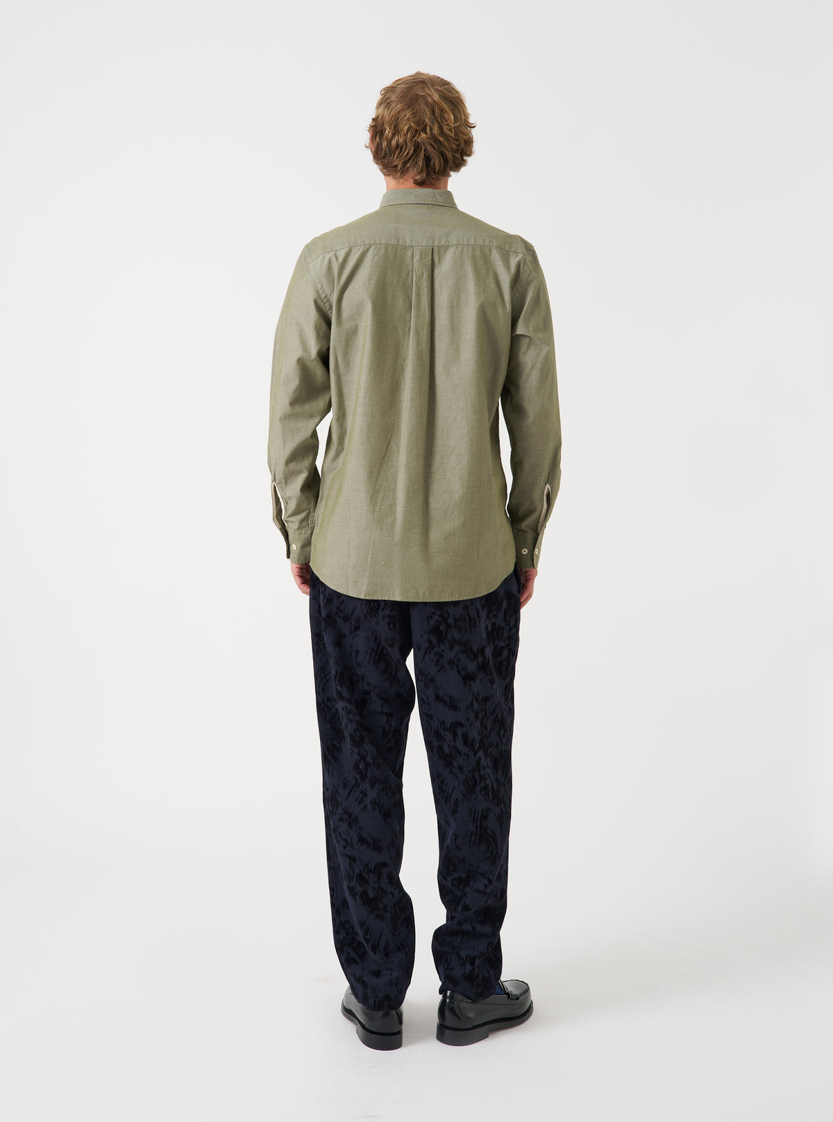 Back view of a male model wearing the Olive Prince Chambray Daybrook Shirt against a light grey studio backdrop. The shirt features a clean back yoke seam, long sleeves, and a relaxed fit with a neat curved hemline.