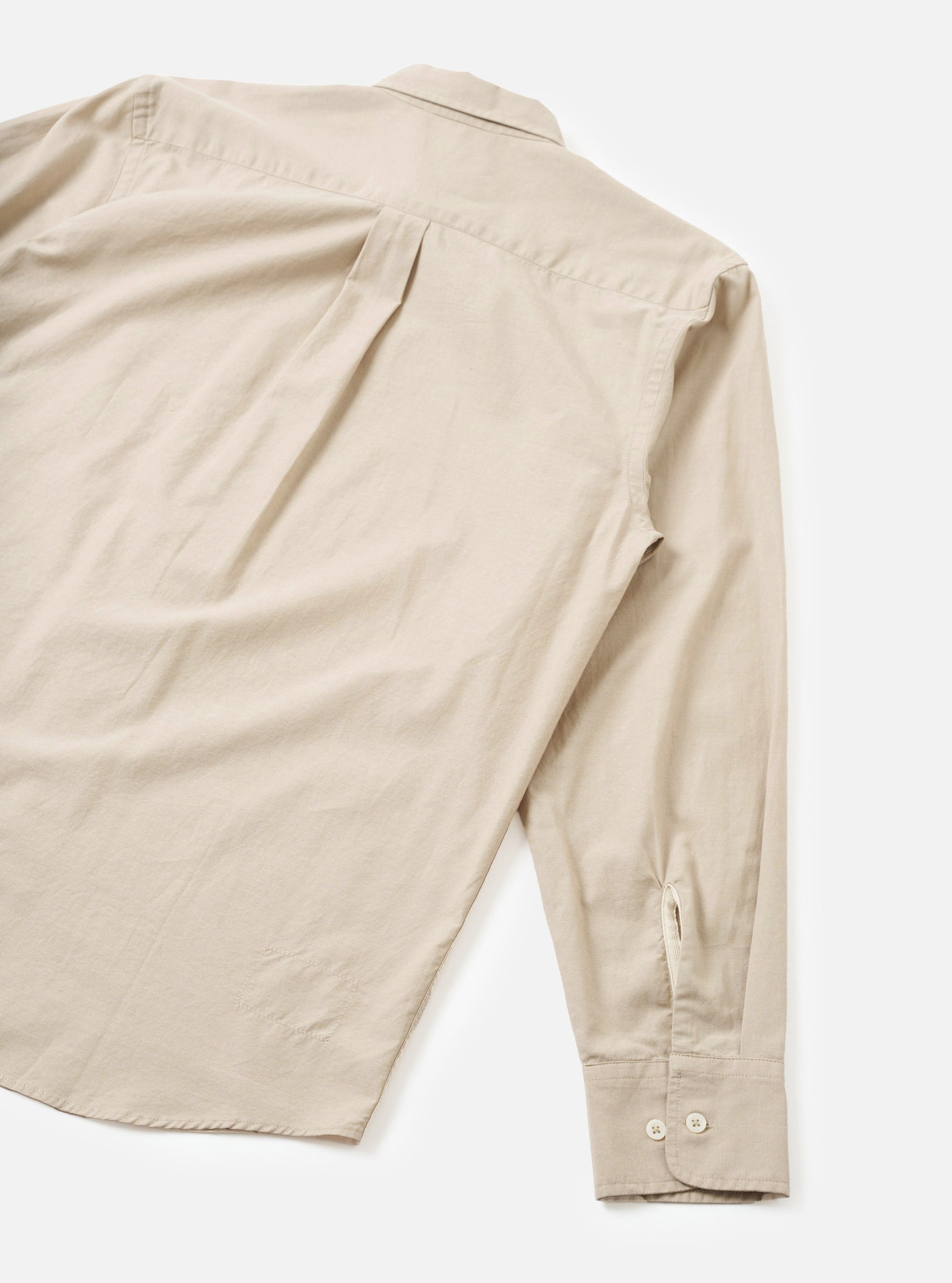 Flat lay of a sand-coloured chambray Daybrook shirt shot from above on a white surface, showing the back and one sleeve. The back features a centre pleat and a small locker loop at the collar. The long sleeve ends in a buttoned cuff with two cream buttons and a placket vent. The spread collar is visible at the top. Fabric has a fine, slightly textured weave.