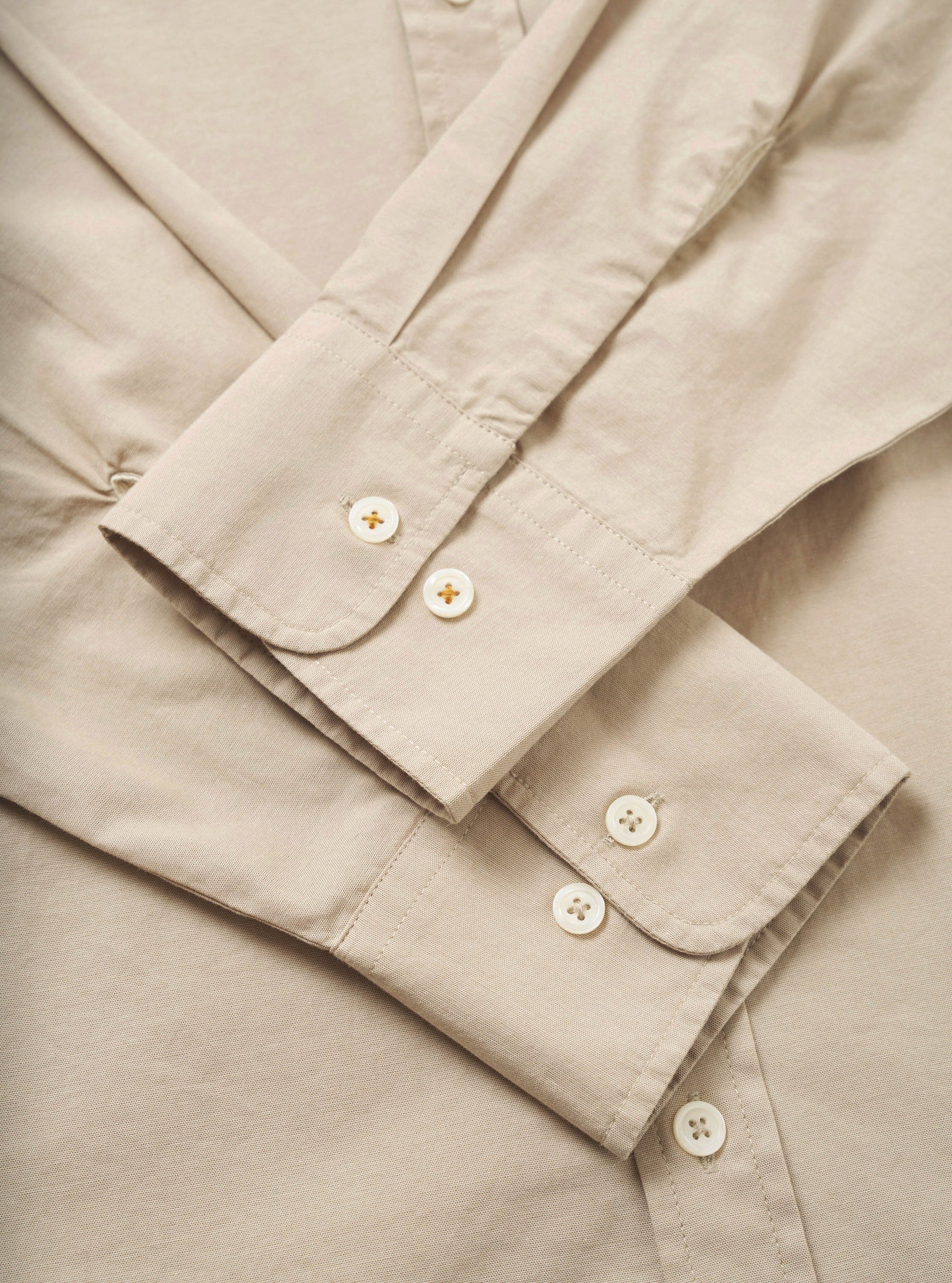 Close-up flat lay of a sand-coloured chambray shirt, focusing on both cuffs. Each single-button cuff features two white mother-of-pearl buttons on a rounded placket with visible topstitching. A placket with a third button is visible at the top of the frame. The plain-weave cotton fabric has a fine, smooth texture.