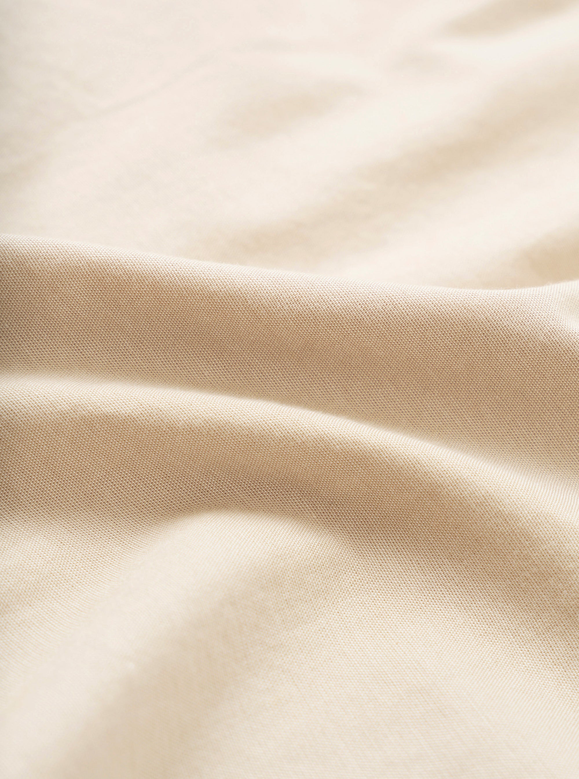 Close-up of the sand-coloured chambray fabric used in the Daybrook Shirt. The fine woven texture is clearly visible, with soft natural folds creating gentle shadow and depth across the cream-toned cloth.