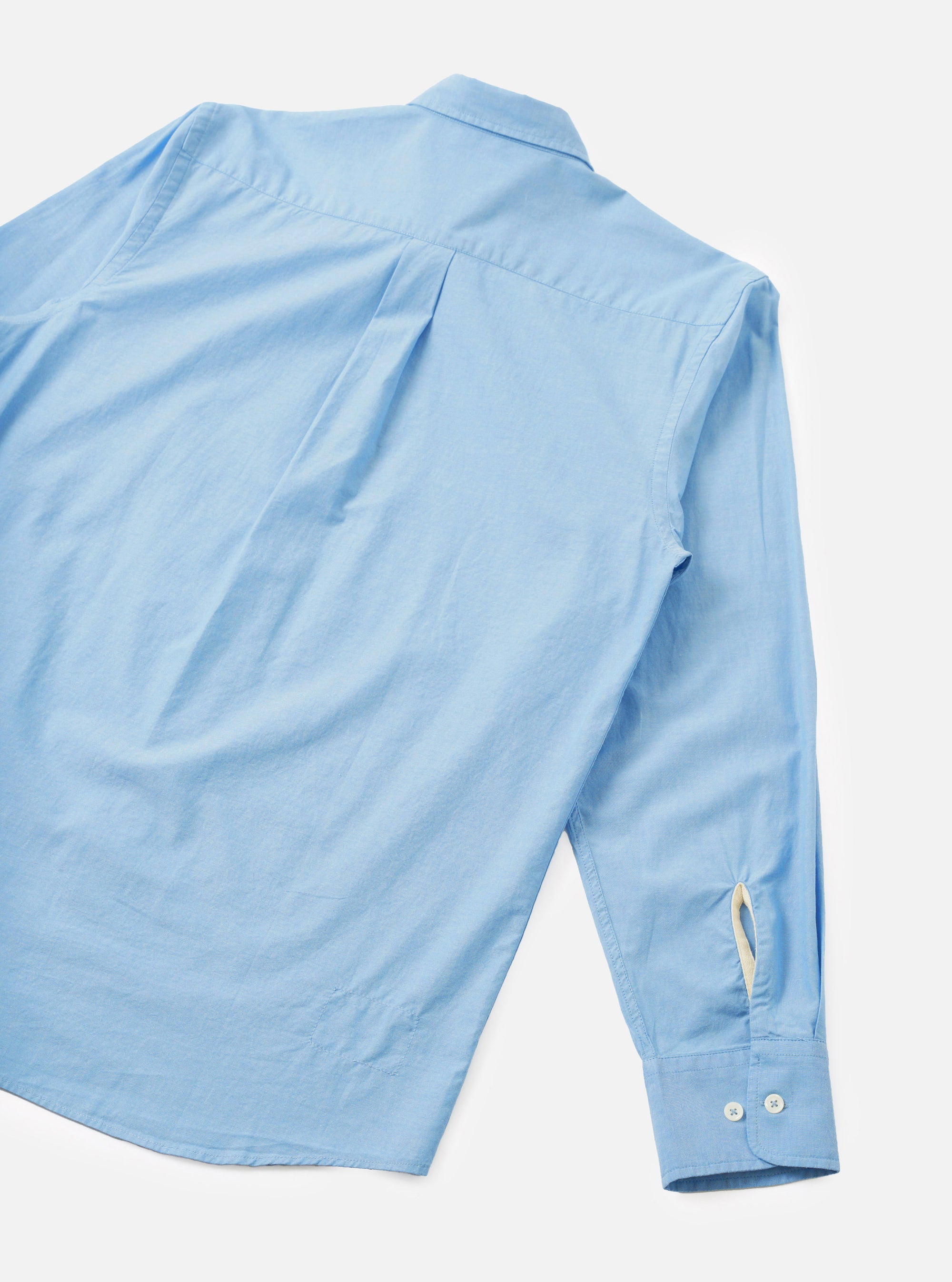 Back view of the sky blue chambray Daybrook shirt laid flat on a white studio backdrop. The plain-weave cotton fabric has a fine, slightly textured finish. Visible details include a back yoke with a central pleat, a spread collar, one long sleeve with a button cuff fastened with two white buttons, and a cream loop tab at the cuff opening.